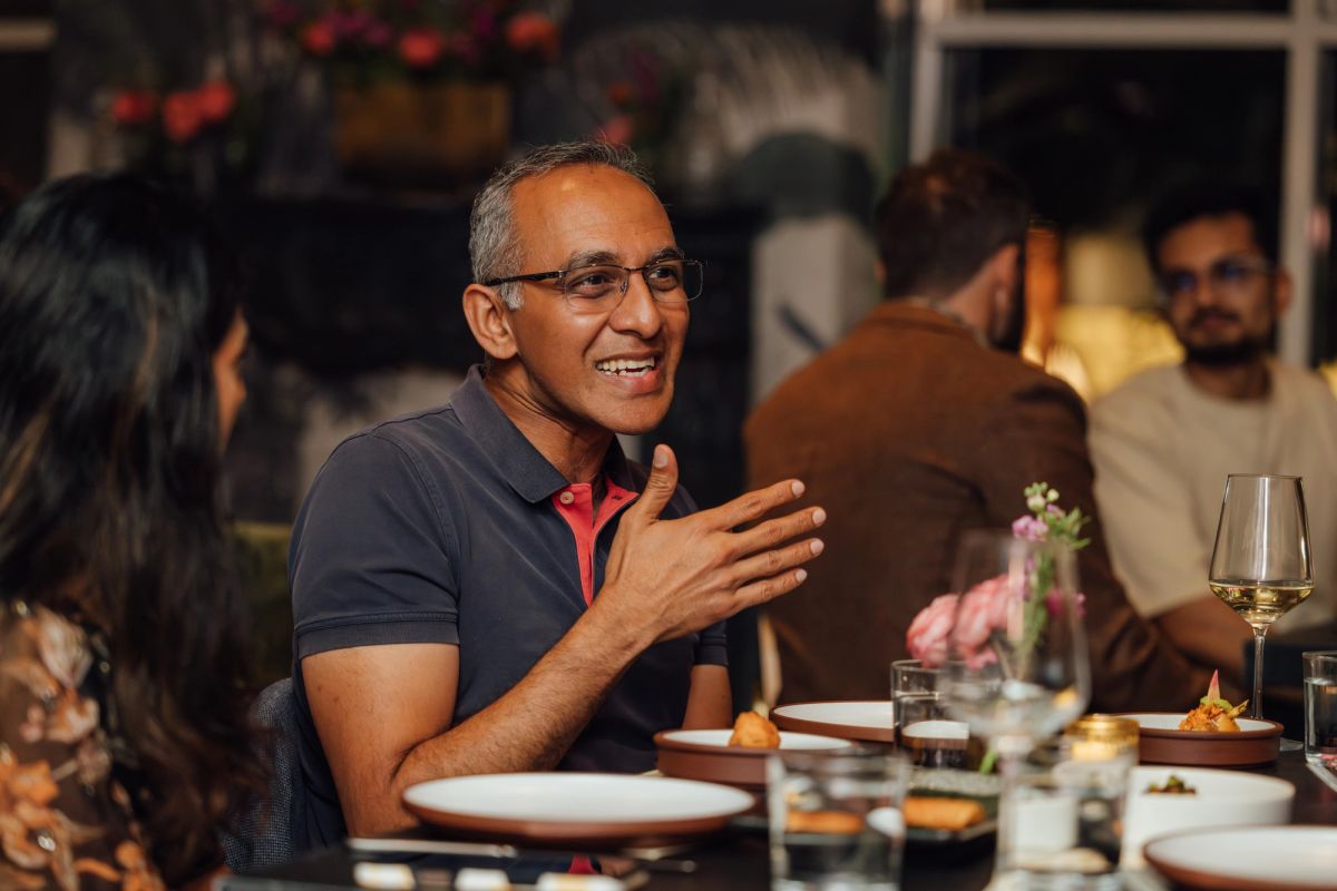 Man in glasses and polo shirt laughing while greeting someone at restaurant table with wine glasses and food