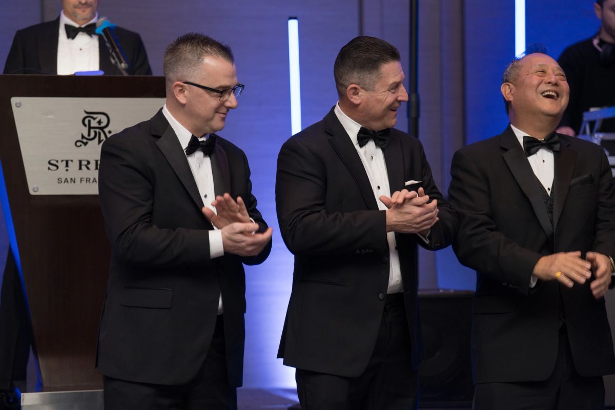 Three men in black tuxedos applauding at a formal gala event, with a podium visible in the background.