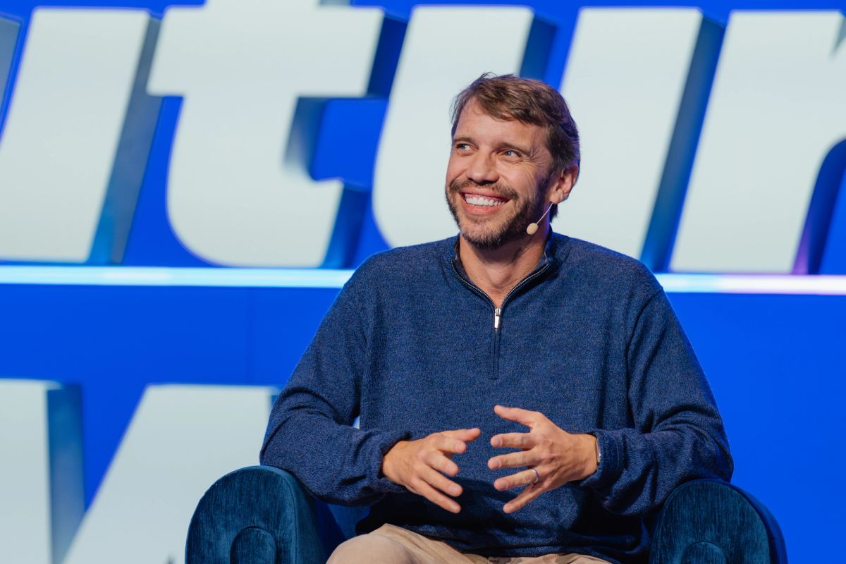 Speaker in blue sweater sitting and gesturing during presentation at conference with blue backdrop