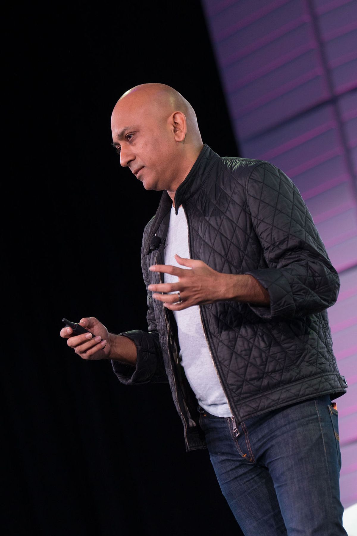 Bald speaker in black quilted jacket gestures while holding phone on stage with purple geometric backdrop