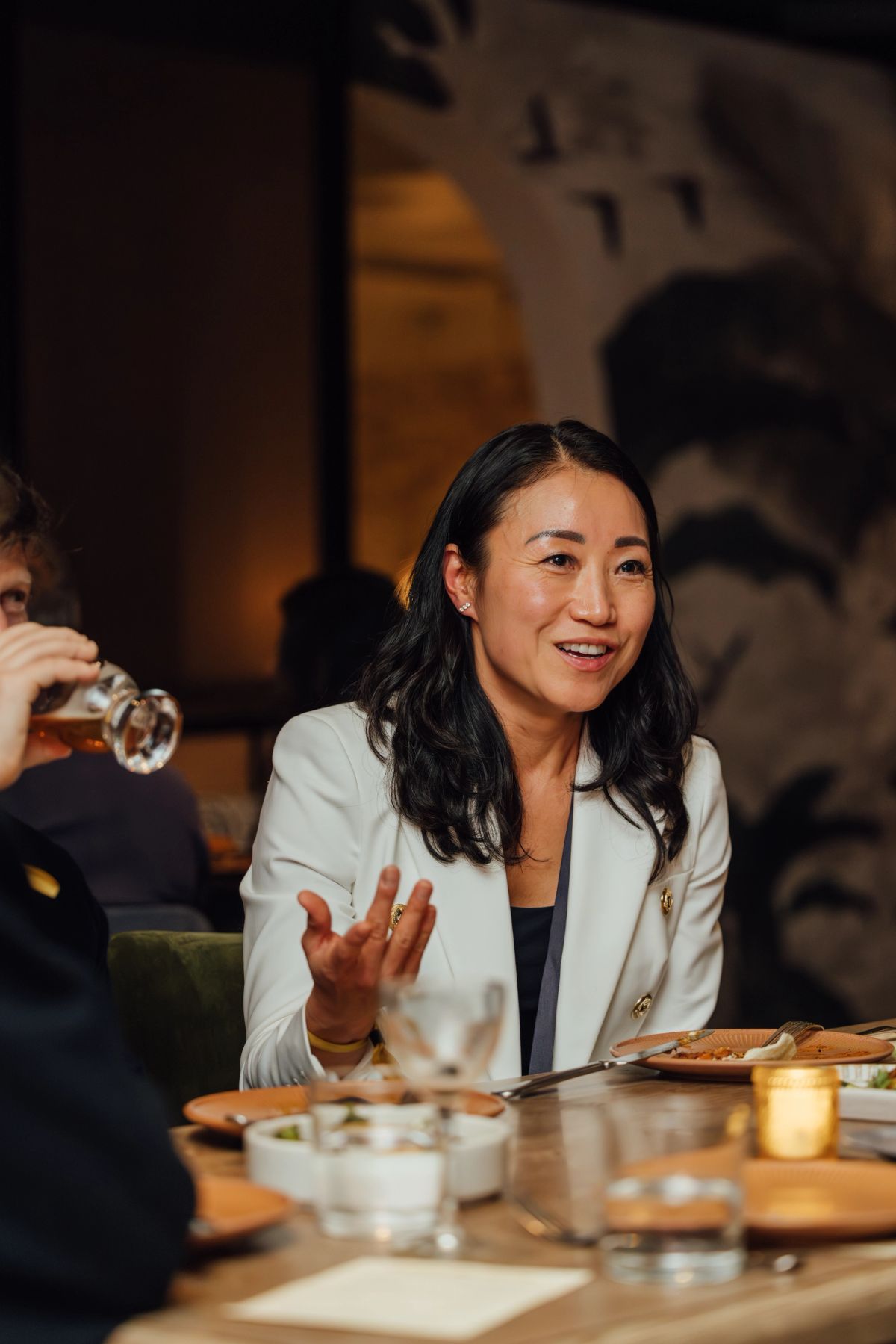 Woman in white blazer gesturing while speaking at dinner table with wine glasses and food in restaurant
