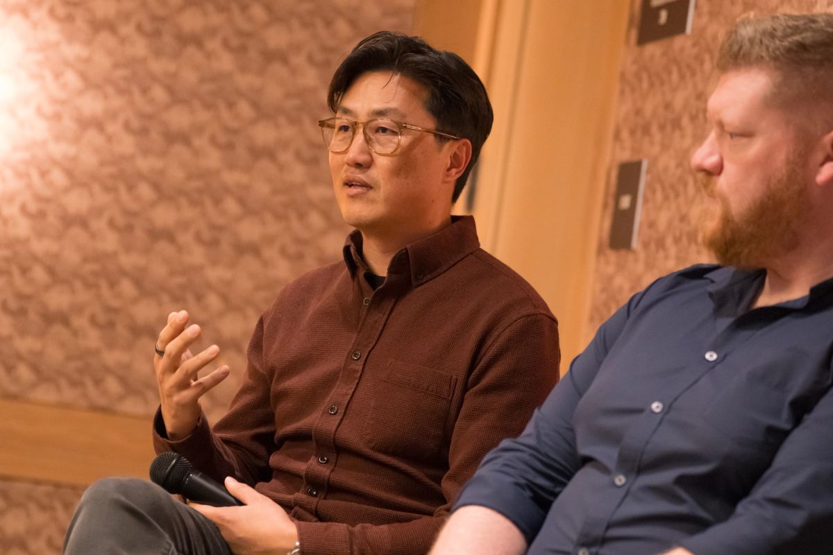 Two men sitting during a panel discussion, one in brown shirt gesturing while speaking, another in blue shirt.
