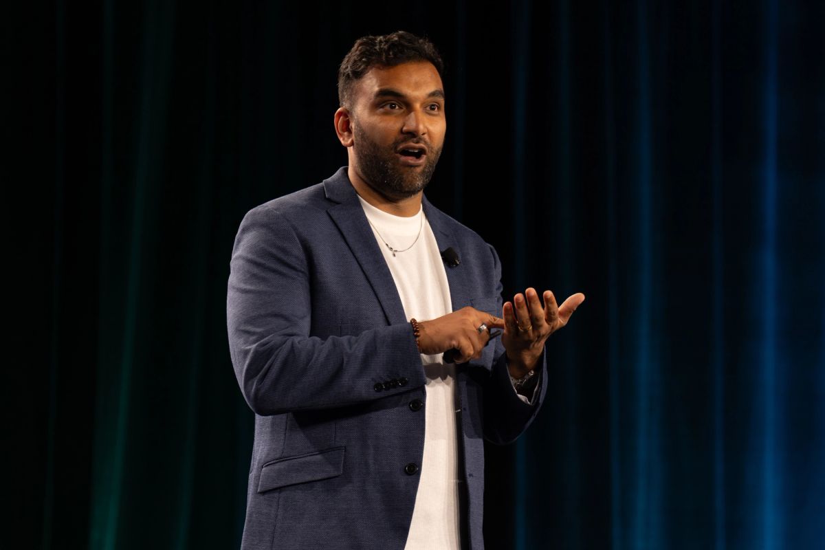 Speaker gestures while presenting on stage, wearing blue blazer and white shirt.