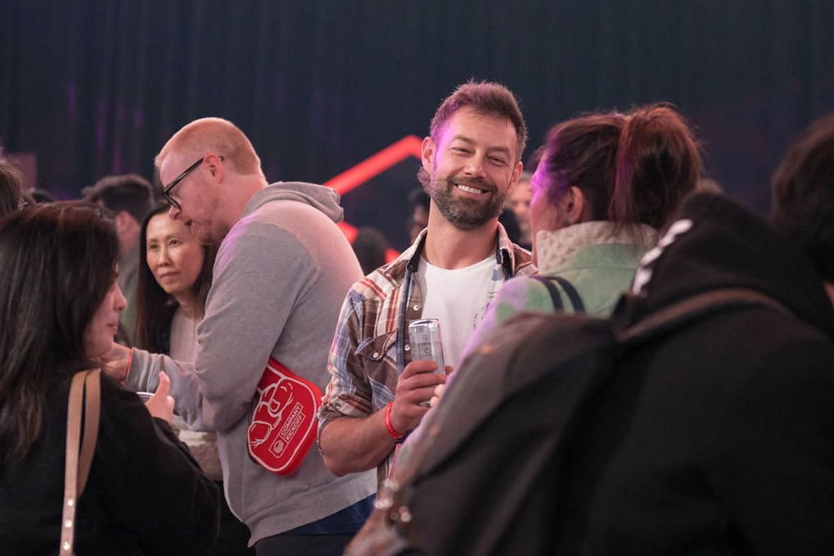 Attendees networking at a conference event, with a bearded man in a plaid shirt smiling while holding a drink