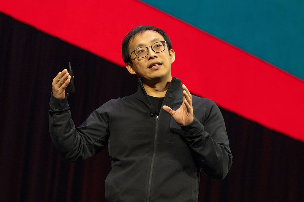 Speaker in black jacket gesturing while presenting on stage with colorful geometric backdrop
