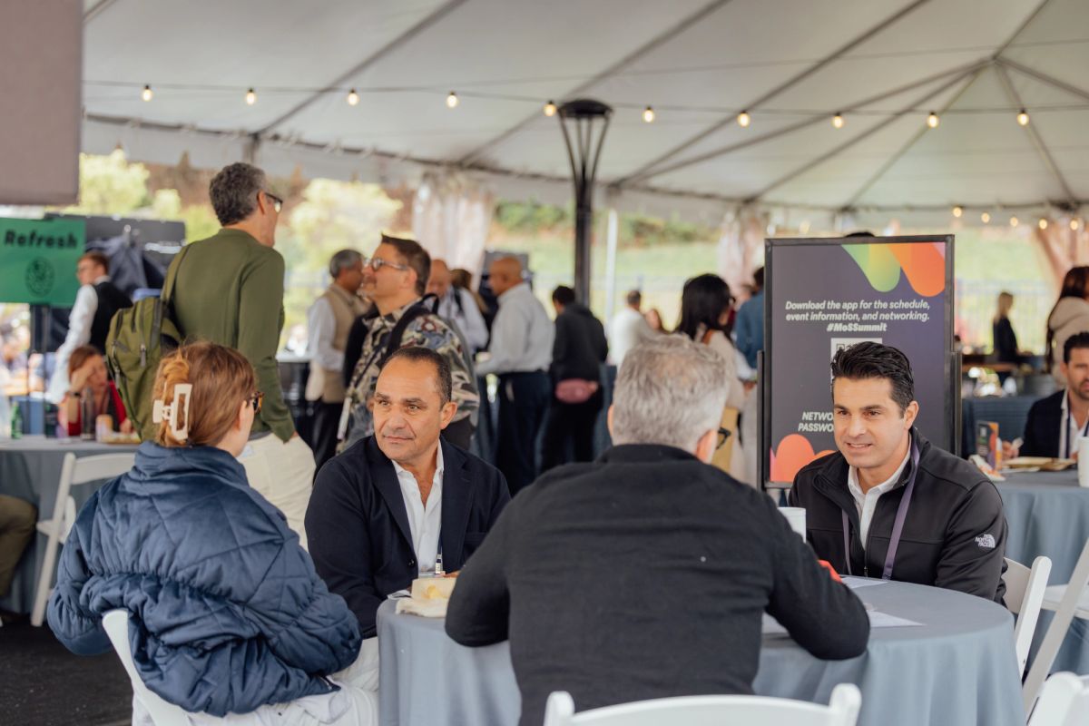 Business professionals networking at tables under a white tent with string lights and promotional banners