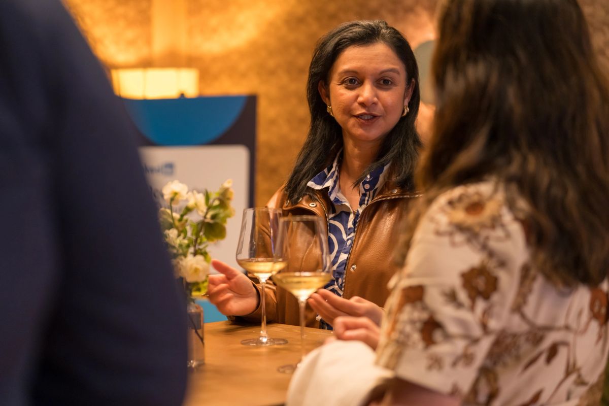 Woman in brown leather jacket holds wine glass while speaking with seated attendees at indoor event