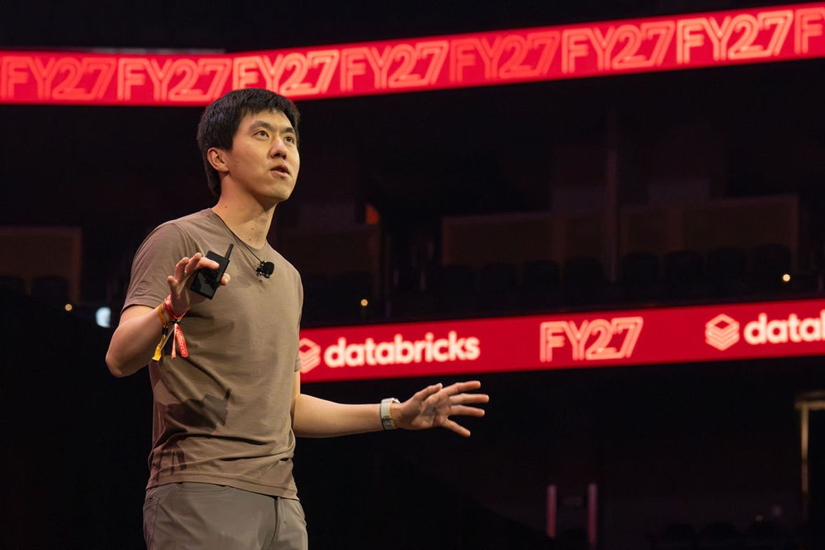 Speaker presenting on stage with microphone at conference venue with red Databricks and FY27 signage