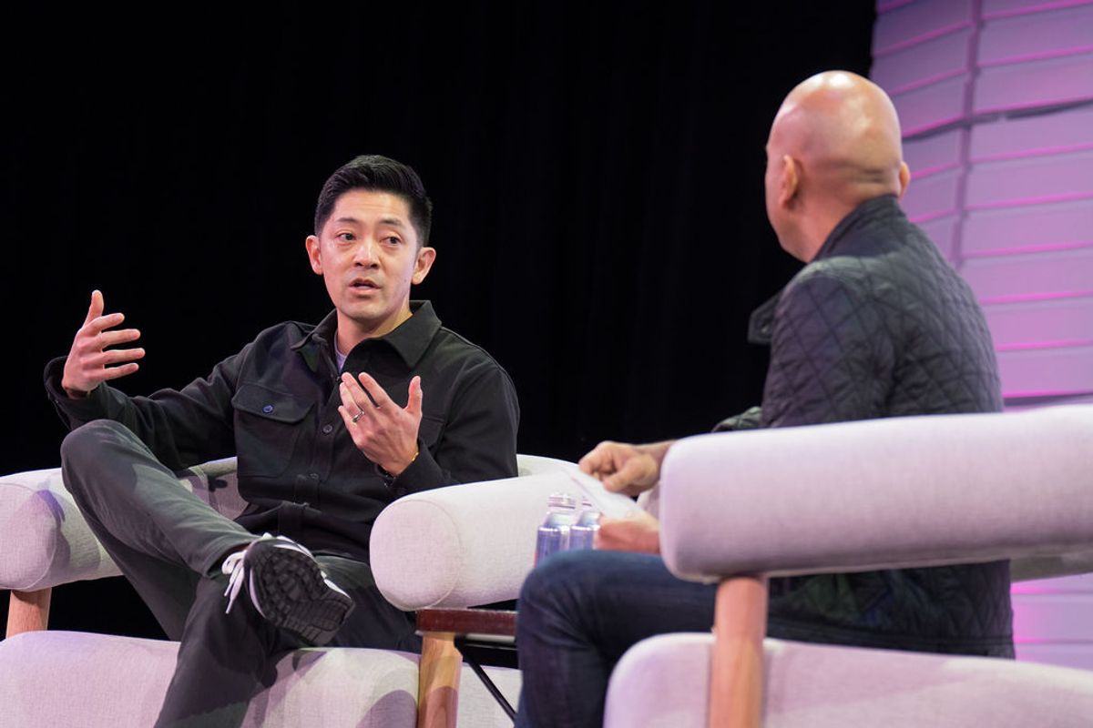 Two men in conversation on stage, one gesturing while speaking, seated in modern white chairs with purple.