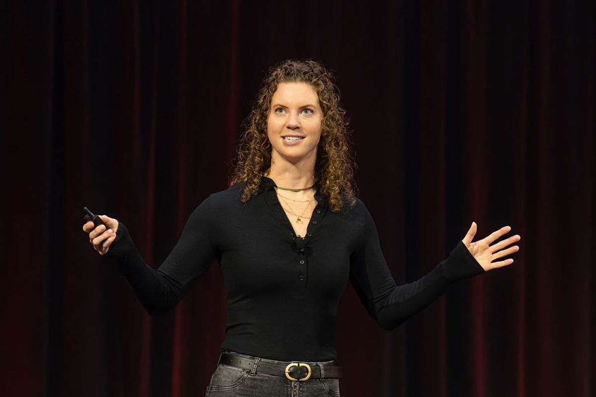 Speaker presenting on stage with microphone, gesturing with one hand while holding device in the other