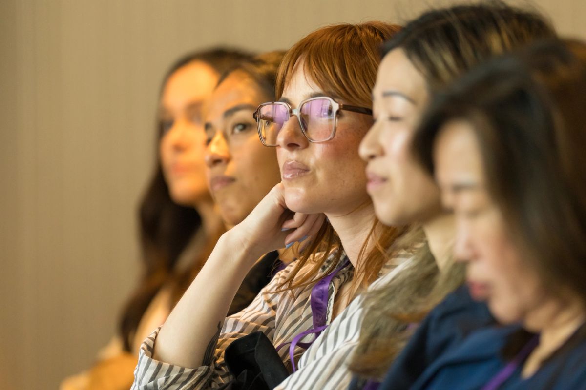 Five women seated in a row listening attentively at a conference session.