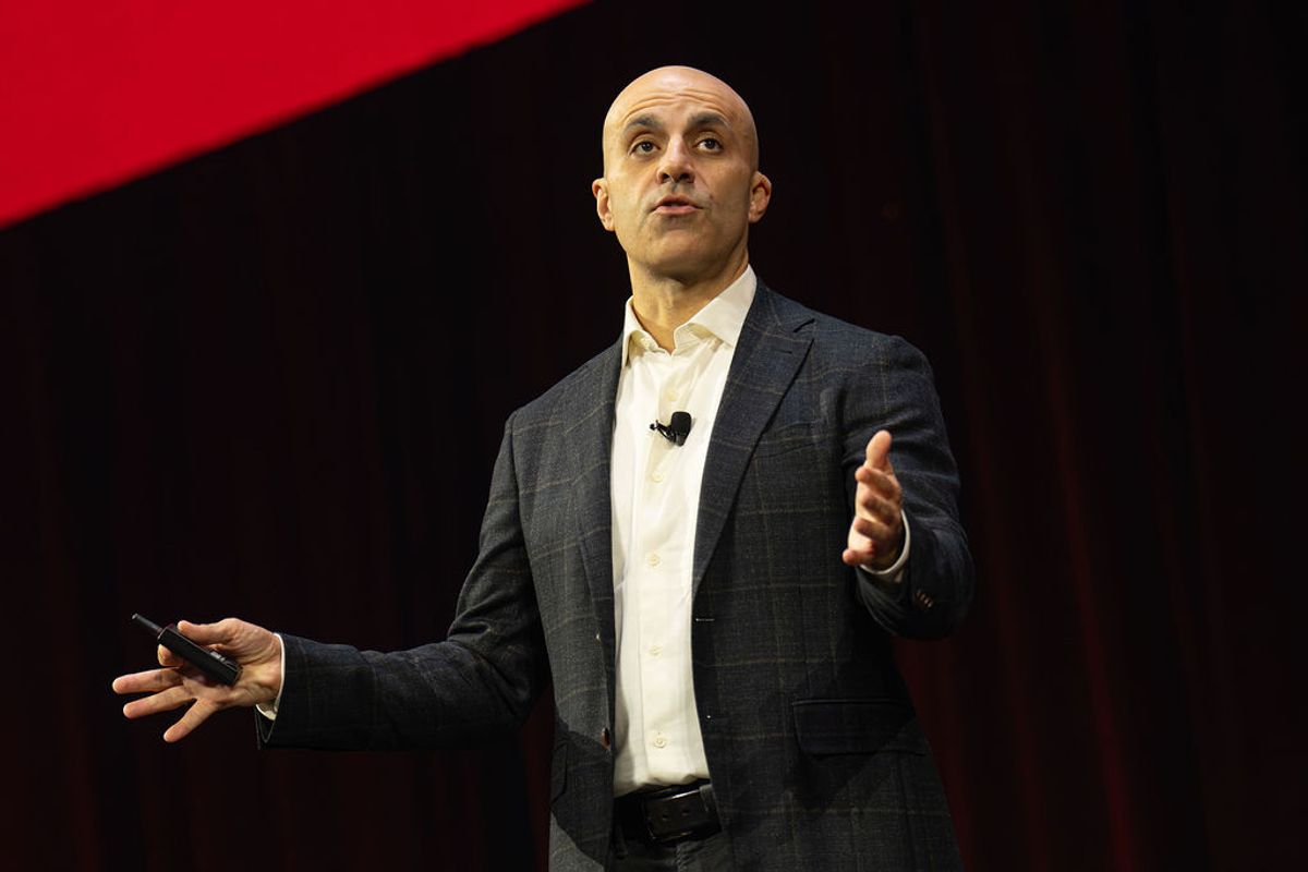 Bald speaker in gray suit presenting on stage with microphone, gesturing while holding device