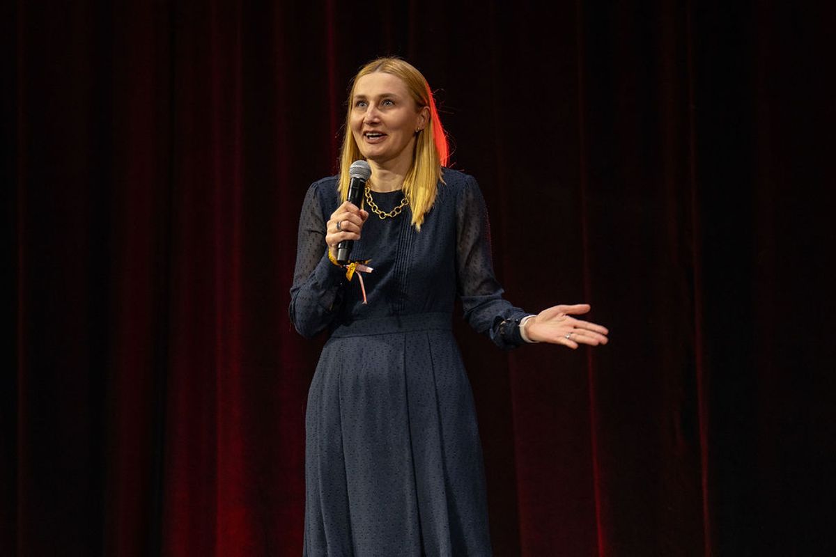 Female speaker in navy dress holding microphone on stage with red curtain backdrop