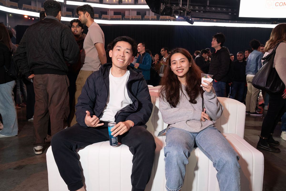 Two attendees sitting on white chairs at a conference event, holding drinks and smiling at the camera