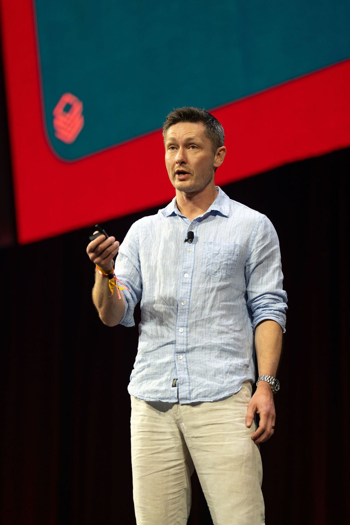 Speaker in blue shirt and khaki pants presenting on stage with red and teal backdrop at conference