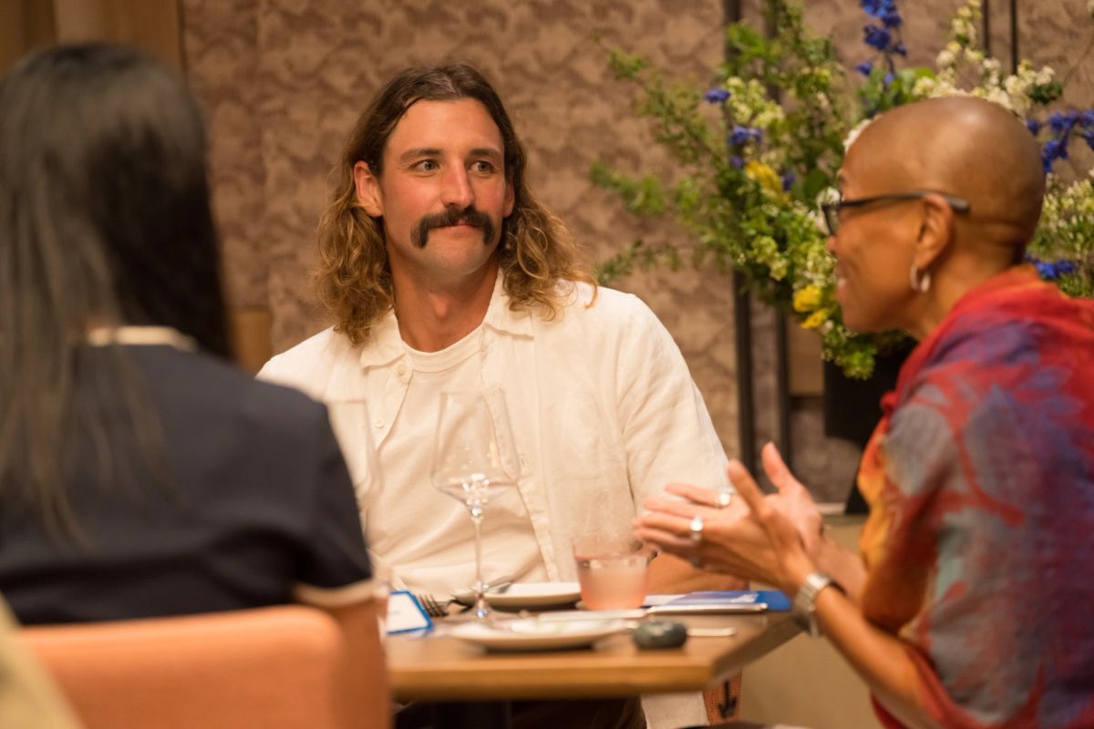 Two panelists sit at a table during discussion, one with long hair and mustache gesturing while speaking
