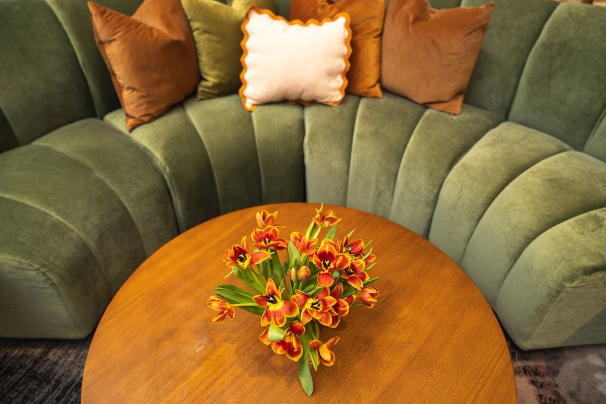 Orange and red tulips in vase on wooden coffee table next to curved green velvet sofa with pillows