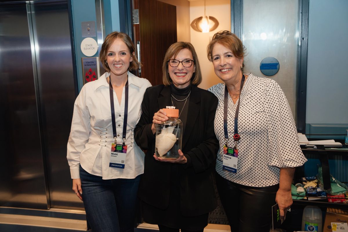 Three women pose together at a conference, with the center person holding an award or trophy near elevator.