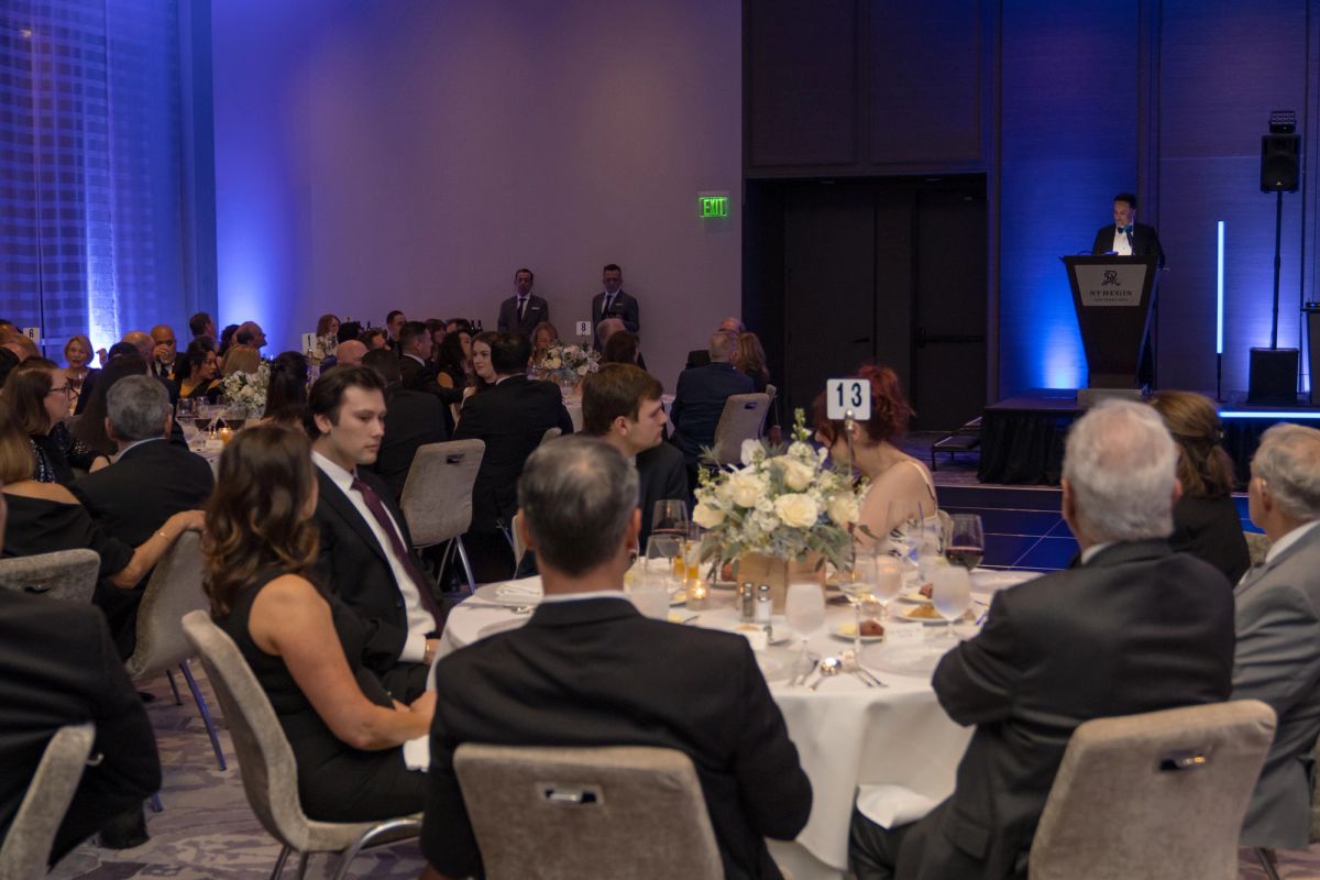 Formal gala dinner with attendees in evening wear seated at round tables while speaker presents at podium