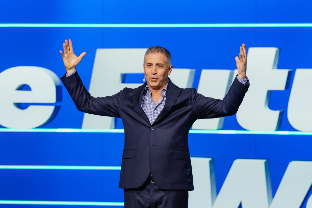 Speaker with raised arms presenting on stage against blue backdrop with white text