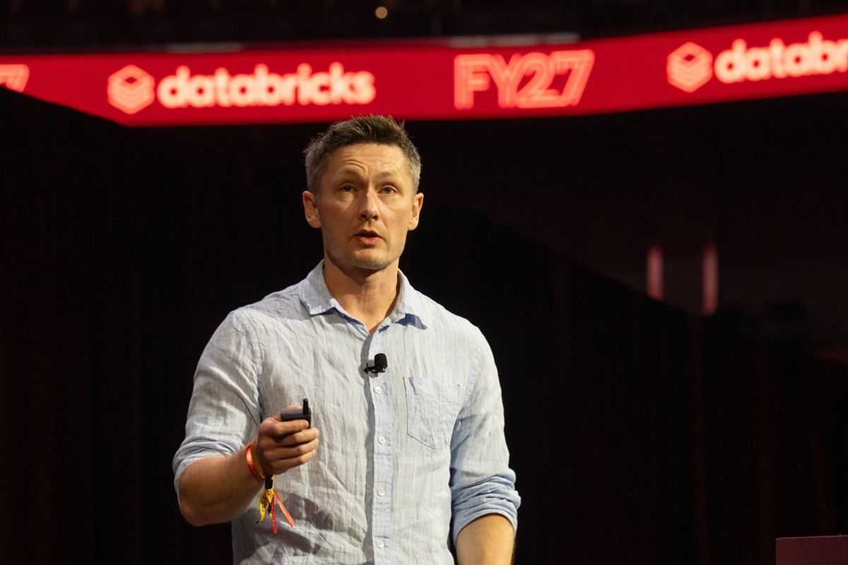 Speaker presenting on stage at Databricks FY27 conference, wearing light blue shirt and holding microphone