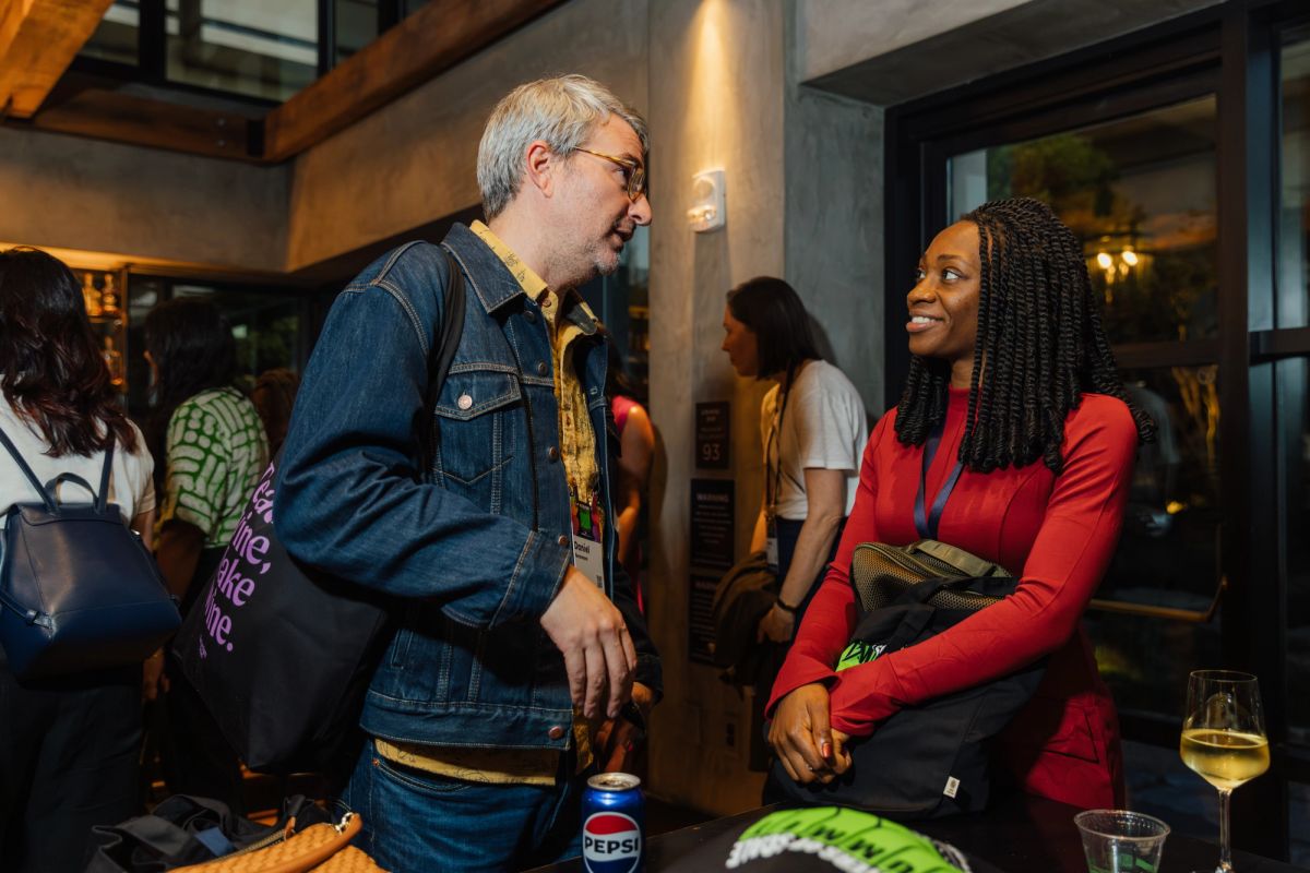 Man in denim jacket and woman in red top having conversation at indoor event with other attendees nearby