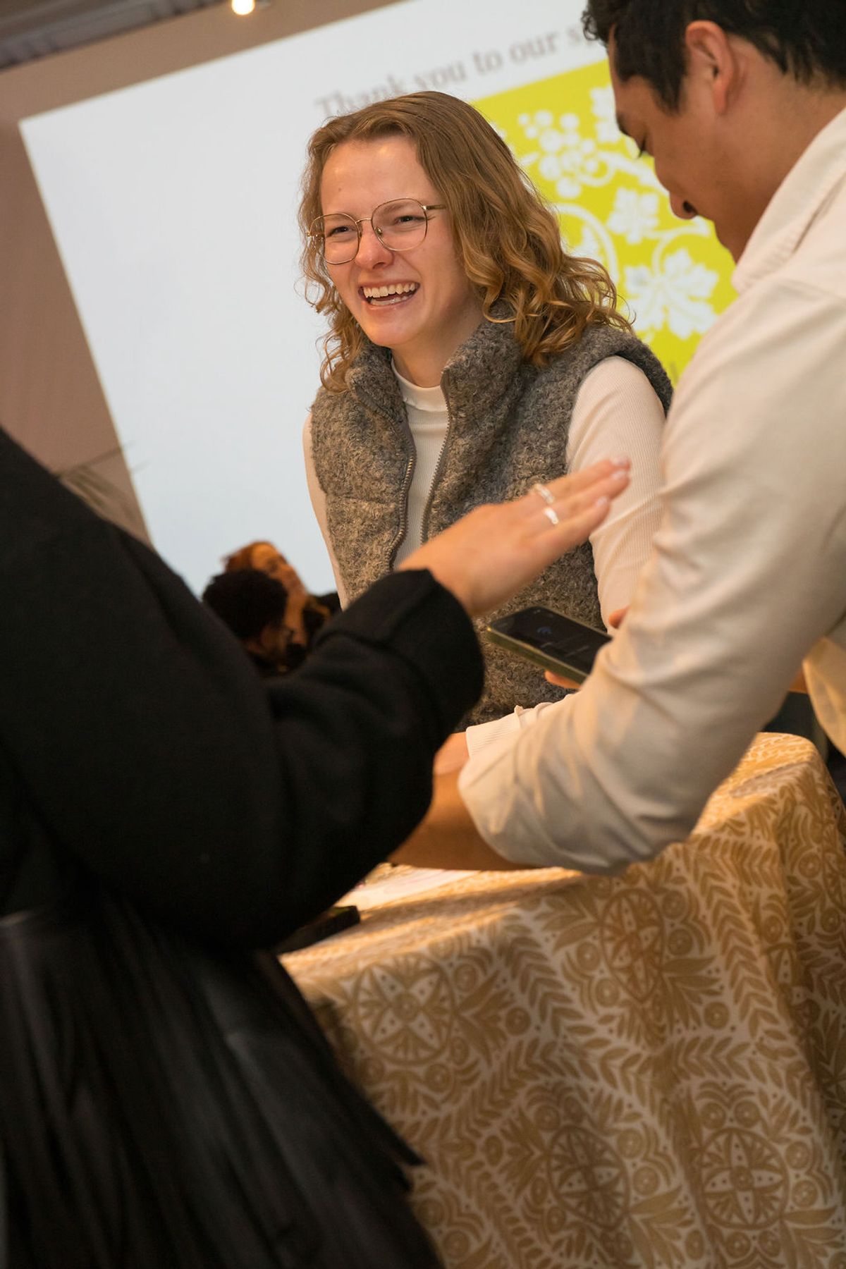 Two people networking at a reception, with a woman in glasses and gray vest laughing while talking to a man.