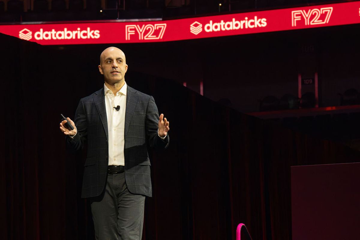 Executive presenting on stage at Databricks FY27 conference with red branded backdrop at Chase Center