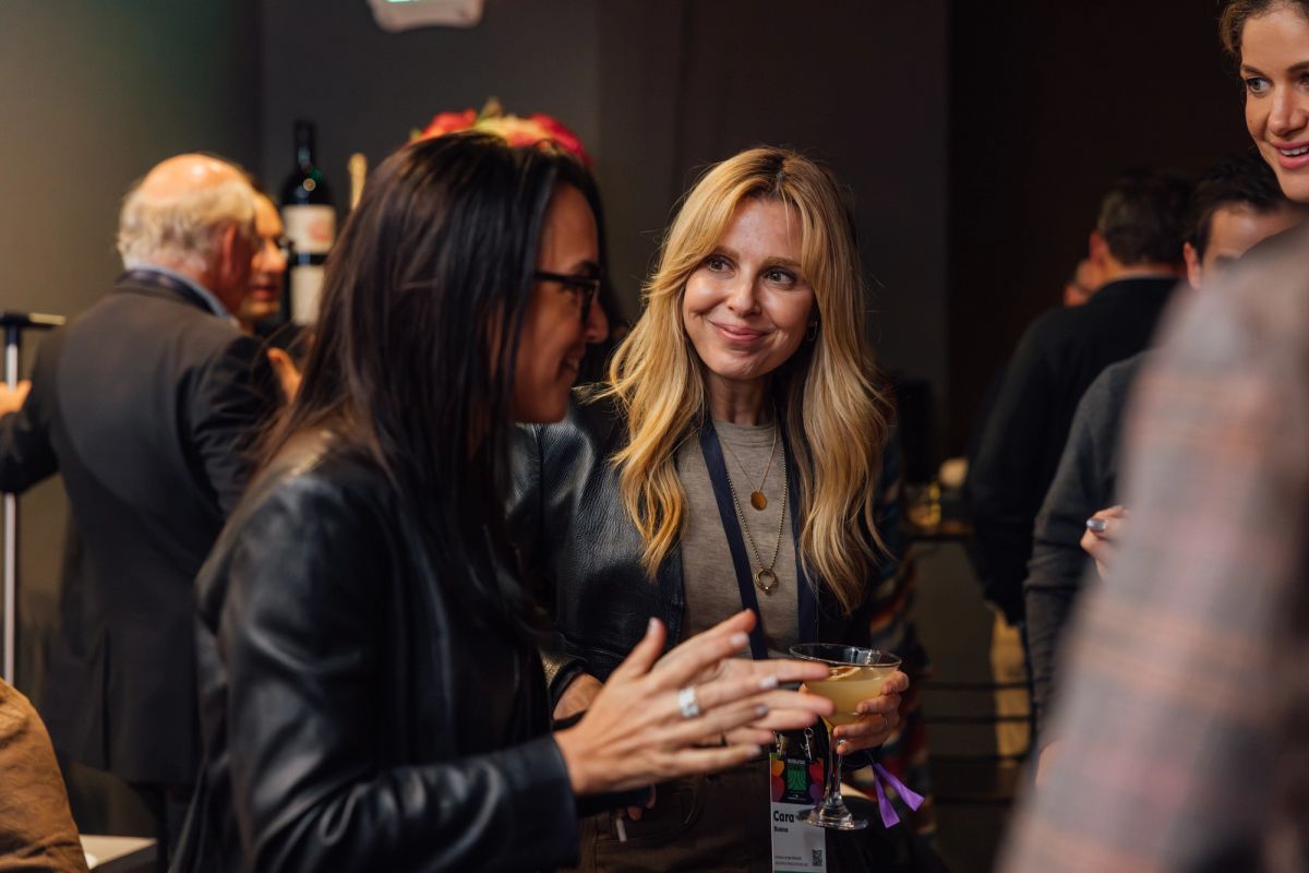 Two women in business attire having a conversation at a networking event with other attendees in background