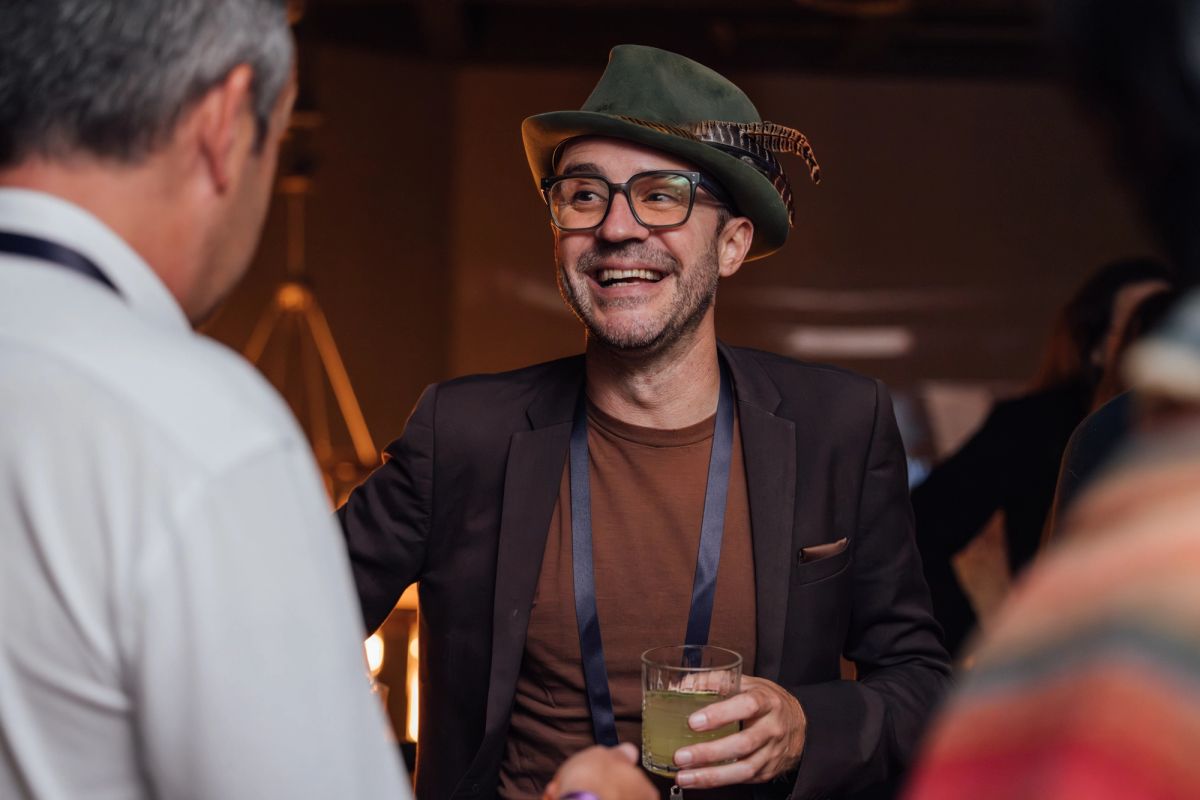 Man in green hat and glasses holding drink talks with another attendee at indoor conference networking event.
