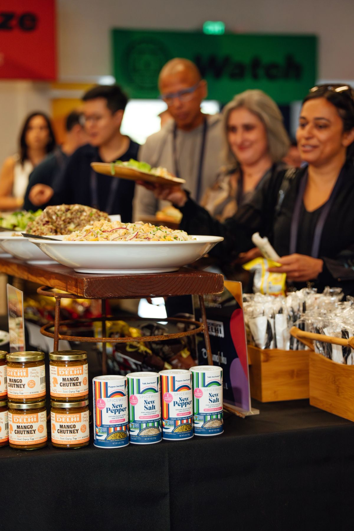 Food sampling station with jars of mango chutney and seasoning products as attendees gather at conference.