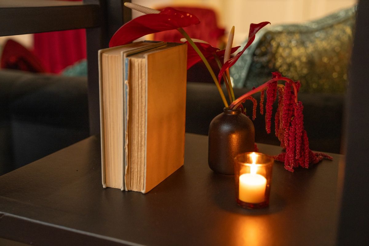 Books, small vase with red flowers, and lit candle arranged on dark wooden table with couch in background