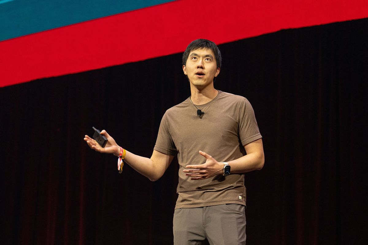 Speaker presenting on stage with brown shirt and jeans, gesturing with hands against dark backdrop with red.