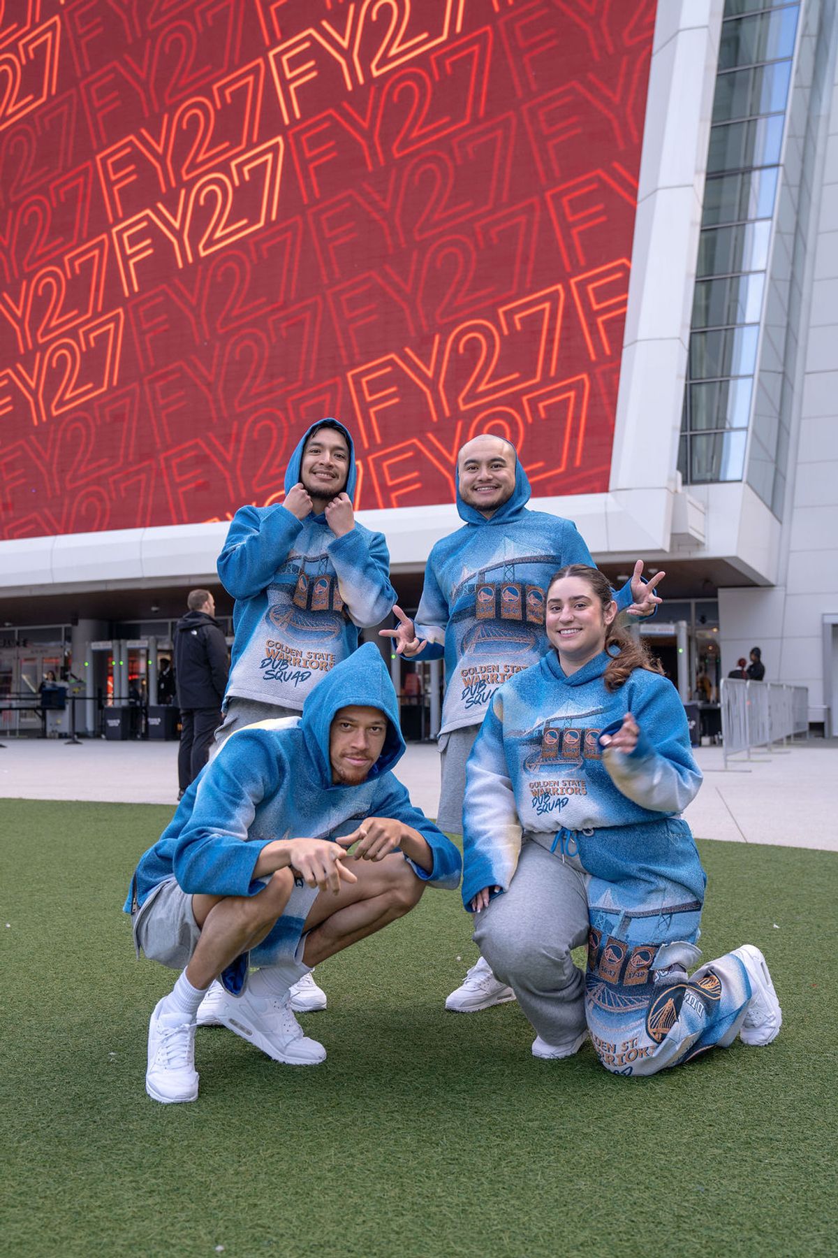 Four people in matching blue hoodies pose together on artificial turf outside a modern building with red.