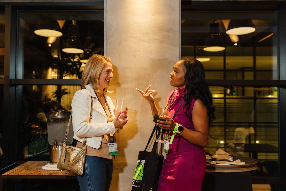 Two women networking and conversing at a conference, one in white blazer, one in pink dress, holding drinks.