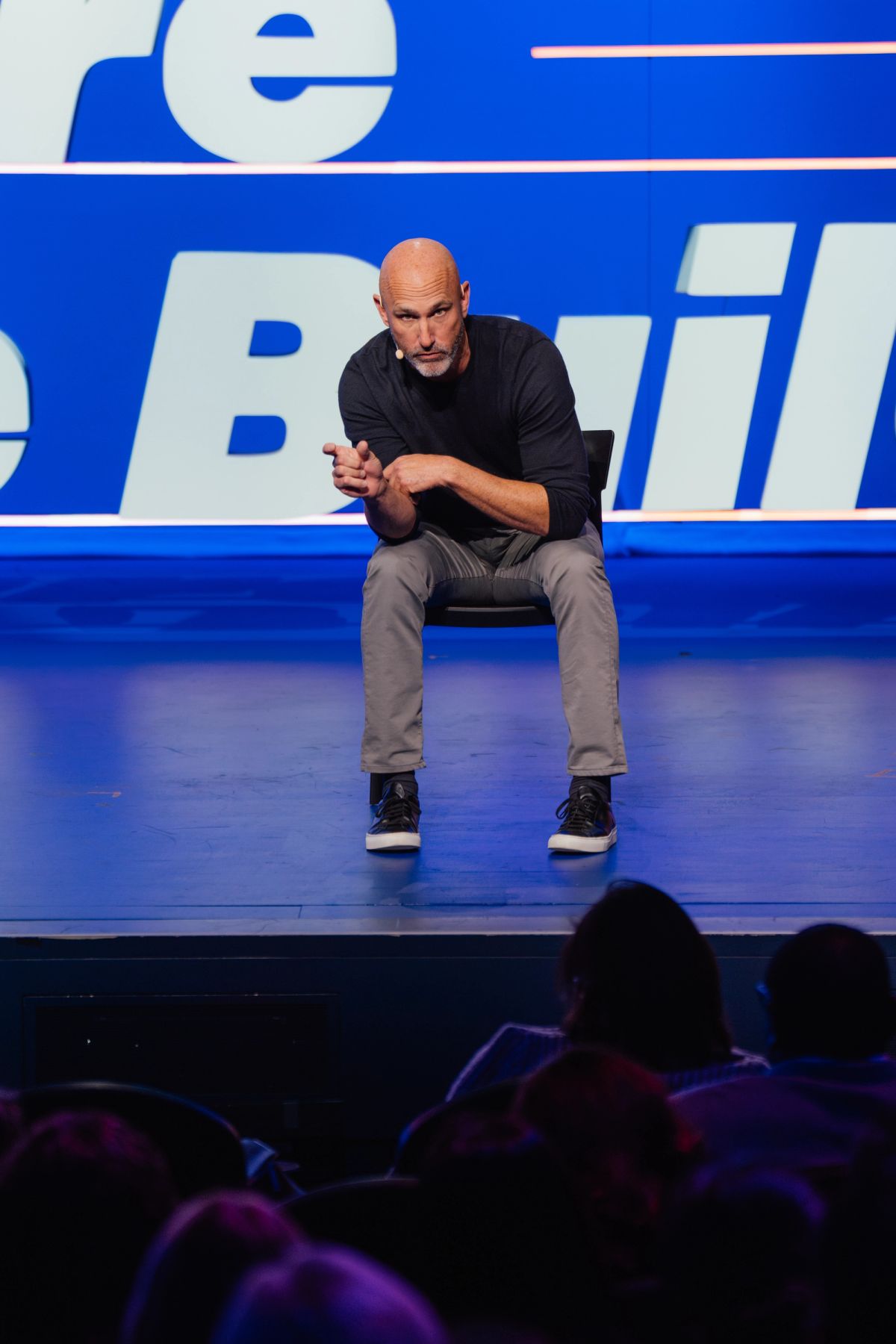 Bald speaker in black shirt sits on chair on blue stage, gesturing to audience below at conference event.