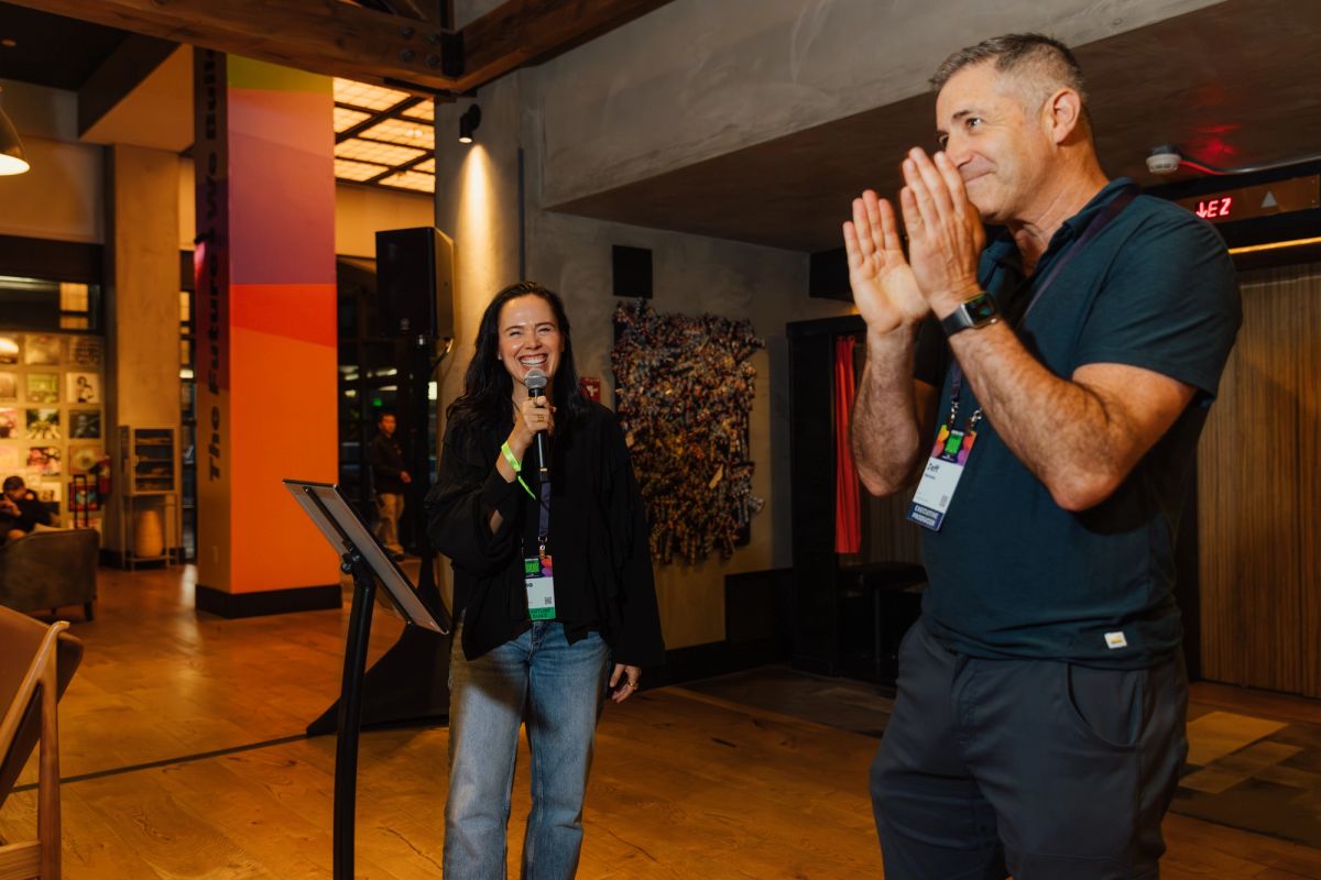 Woman speaking into microphone while man applauds at conference event in modern venue with orange walls