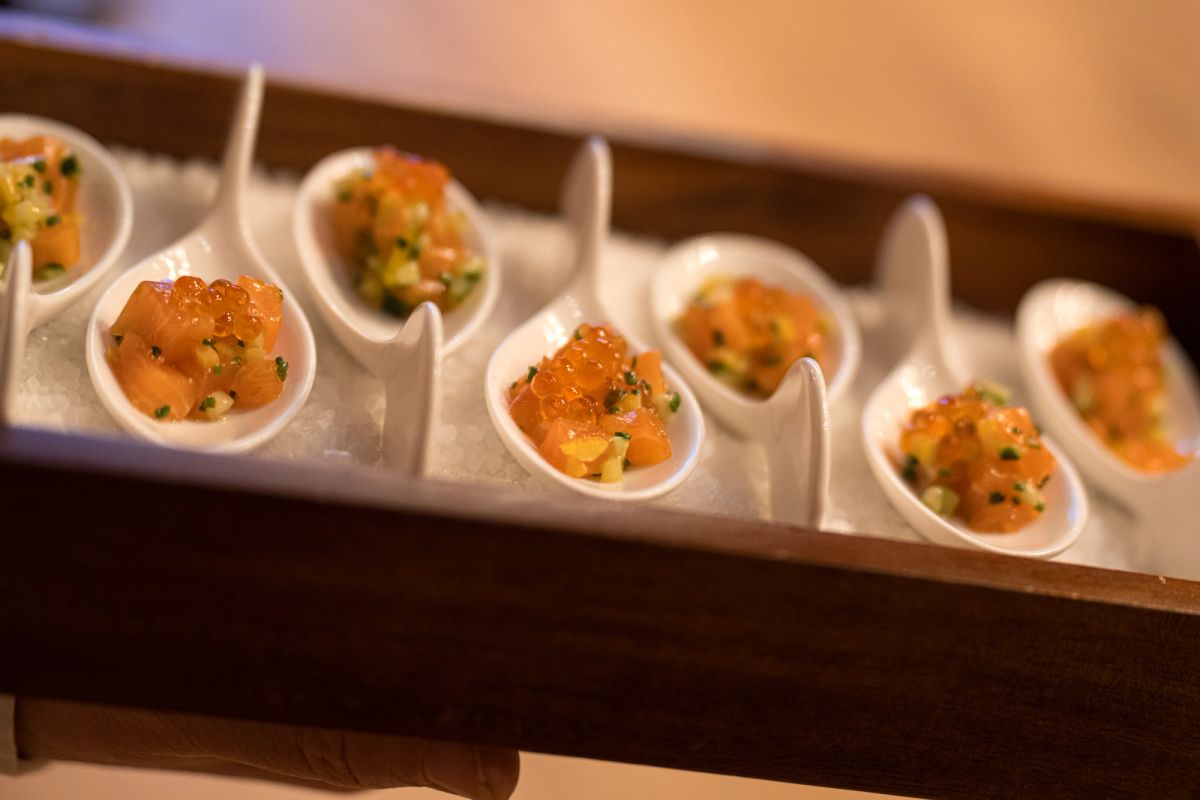 Small white spoons arranged in a wooden tray, each containing orange garnished appetizers with herbs