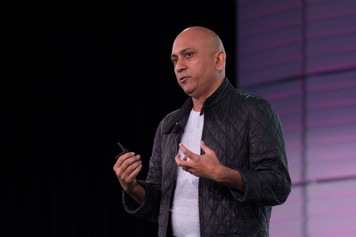 Bald speaker in black quilted jacket presents on stage with purple backdrop, gesturing while holding.
