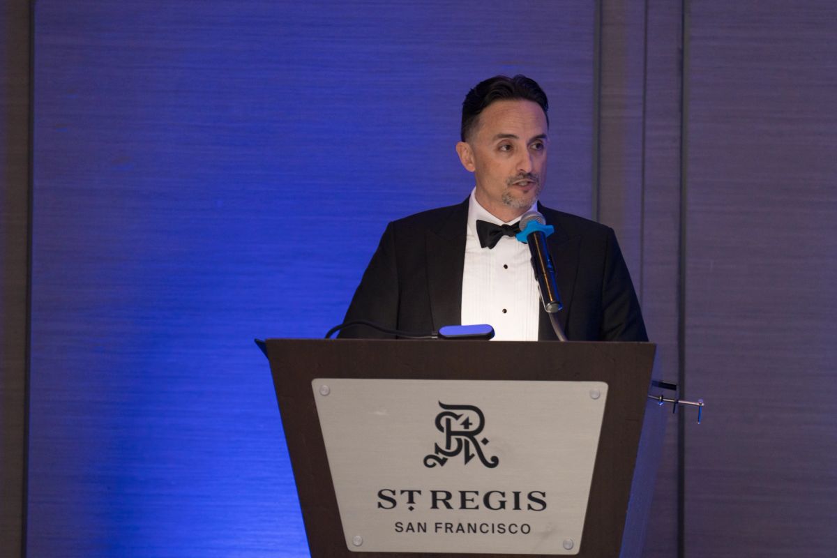 Speaker in tuxedo at podium with microphone at St. Regis San Francisco, blue lighting in background