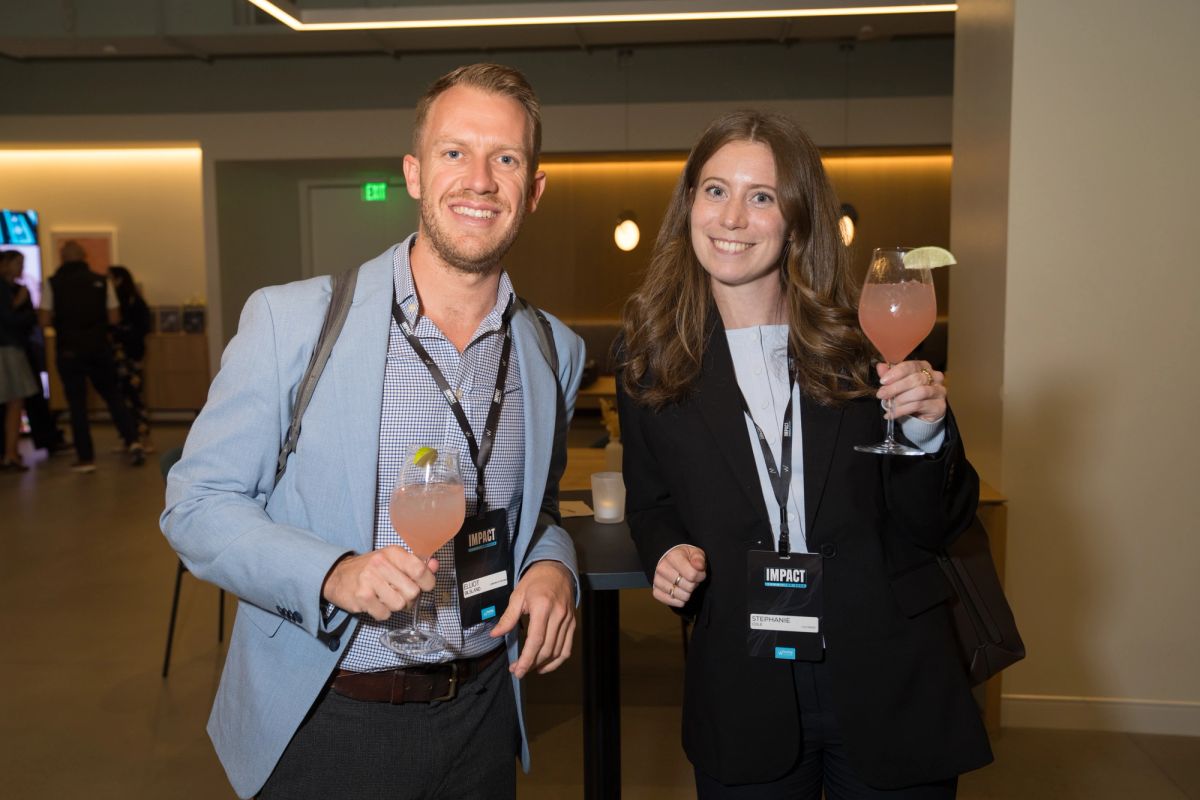 Two conference attendees holding cocktails and wearing lanyards smile in a modern venue lobby