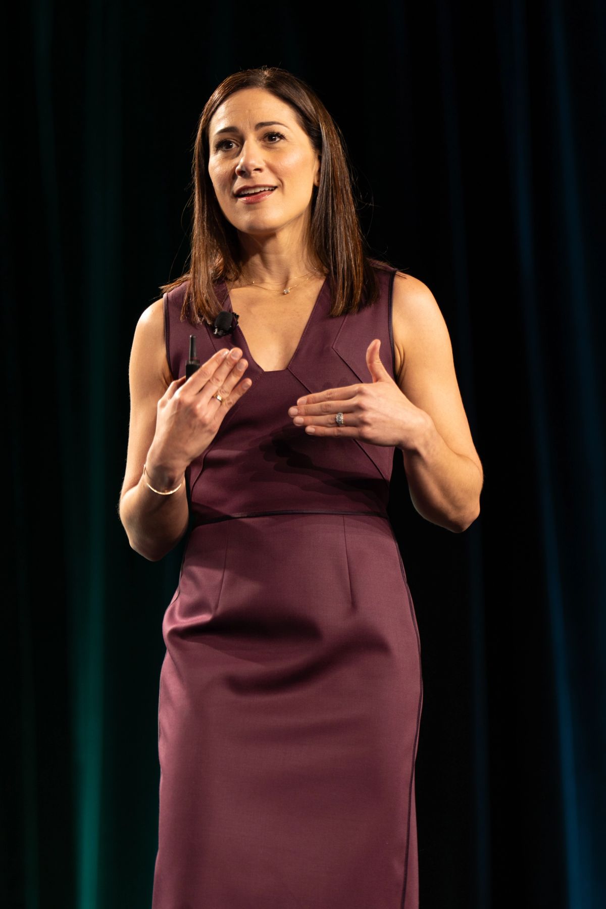 Woman in burgundy dress gestures while speaking on stage before dark curtain backdrop.