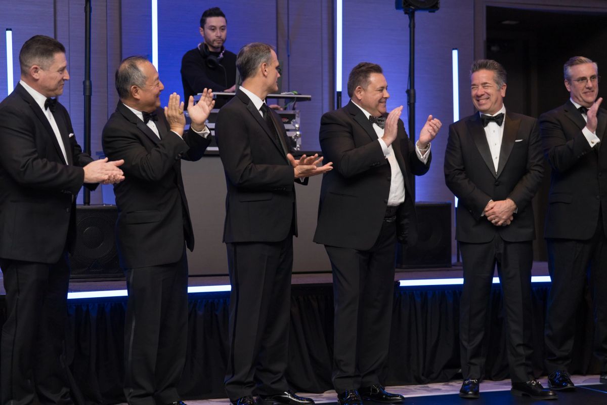 Seven men in black tuxedos applauding on a stage with purple and blue lighting at a formal gala event.