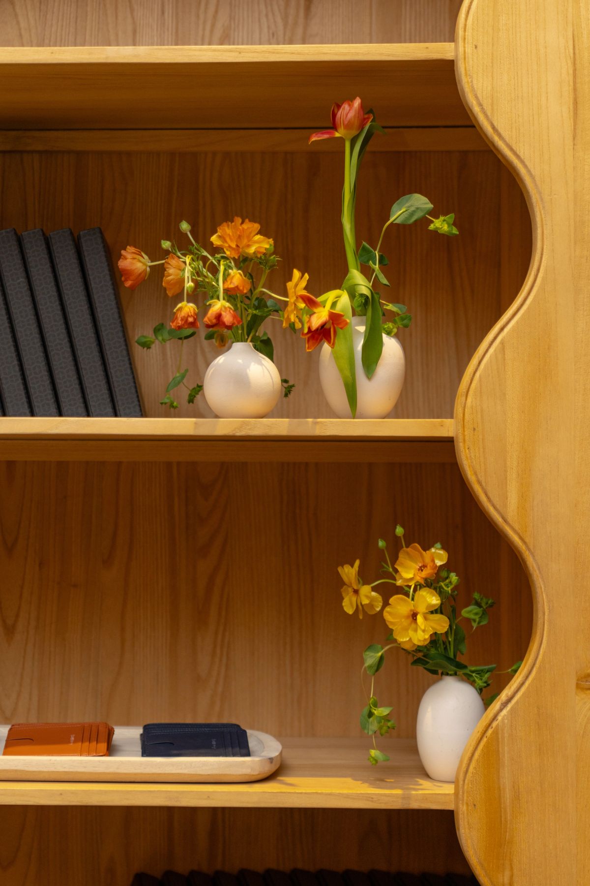 Wooden shelving unit with white ceramic vases holding orange and yellow flowers, books, and decorative items