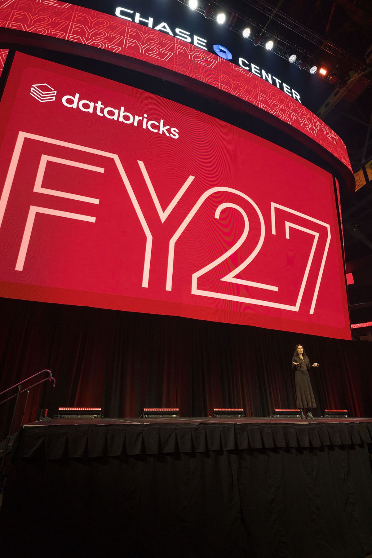 Speaker presenting on stage at Databricks FY27 conference at Chase Center with large red branded screen.