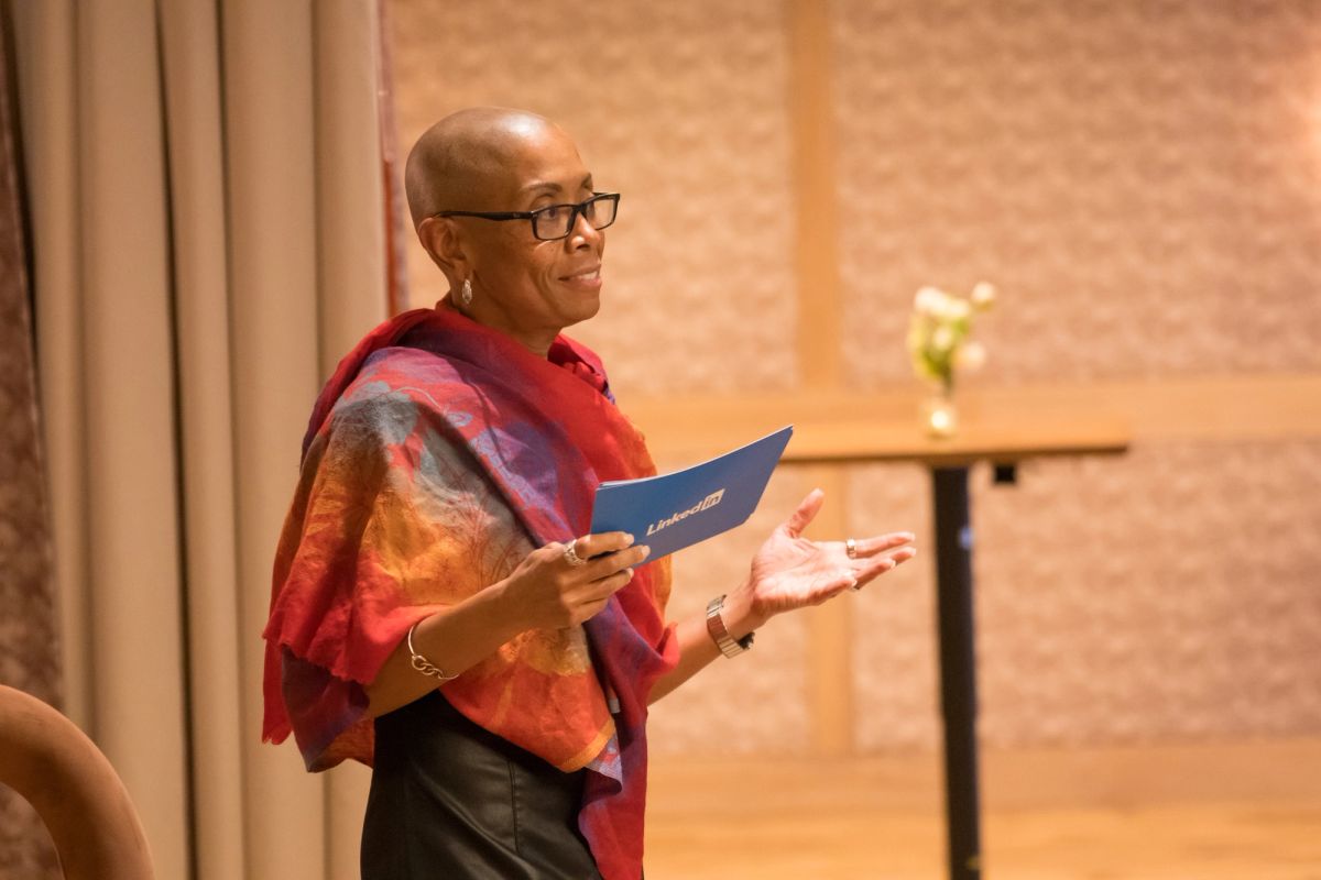 Speaker in colorful shawl holds blue paper while presenting at panel discussion in warm-lit venue