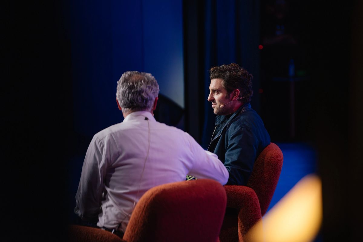 Two people in conversation on red chairs at a conference, one facing away in white shirt, another in dark.