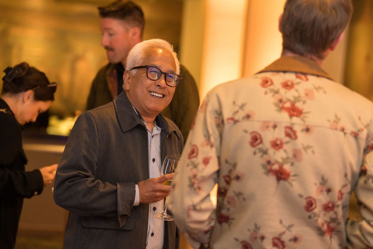 Older man in glasses and blazer holds wine glass while talking to another attendee at networking event
