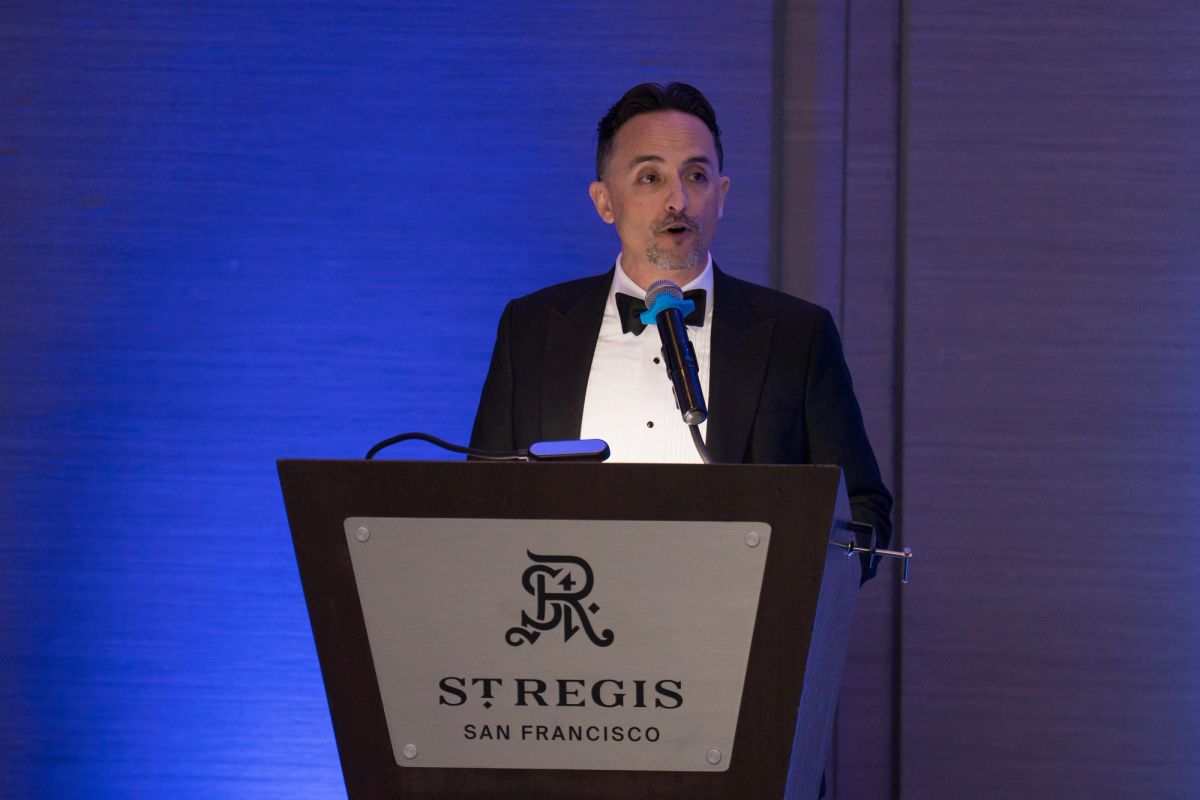 Speaker in black tuxedo presenting at podium with St. Regis San Francisco logo at formal gala event