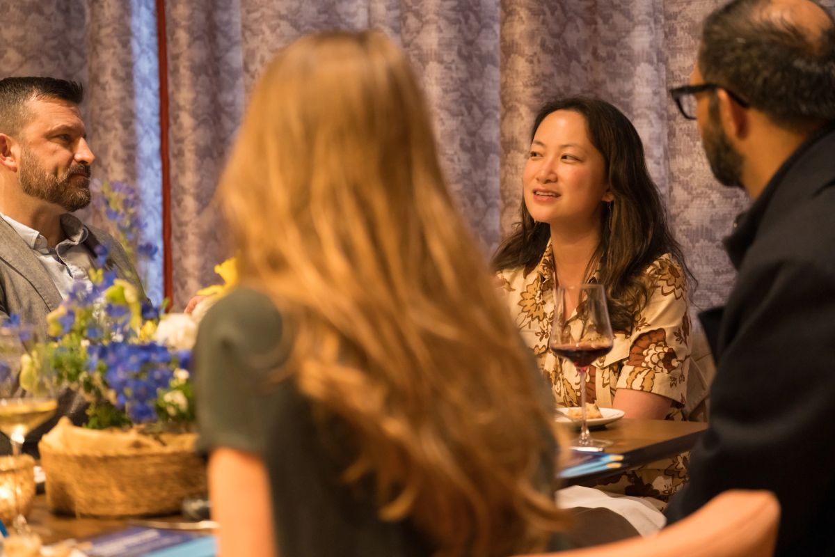 Panel speakers and attendees in discussion at indoor event with floral arrangements and curtained backdrop